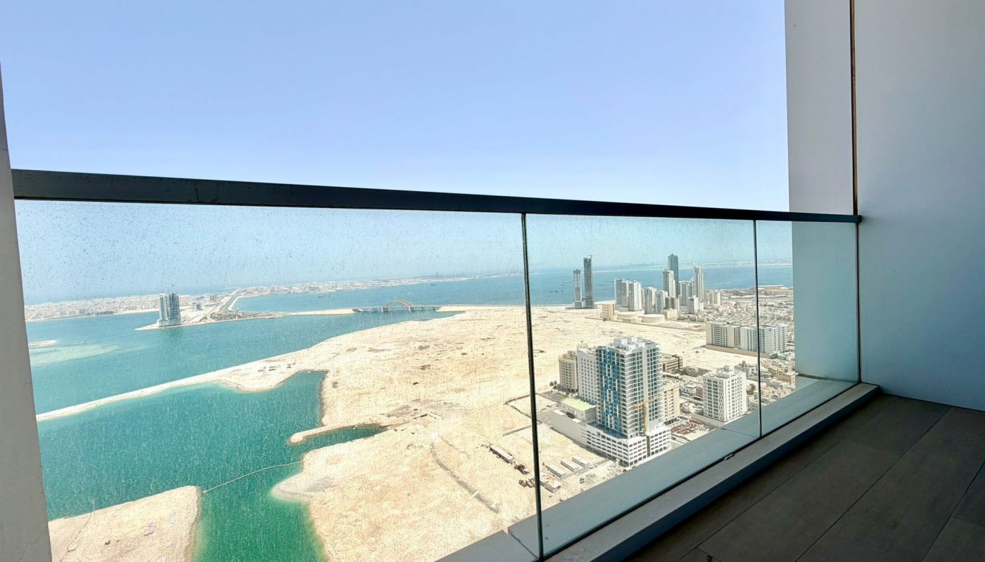 A high-rise balcony with glass railing overlooks a coastal cityscape and water under a clear blue sky.