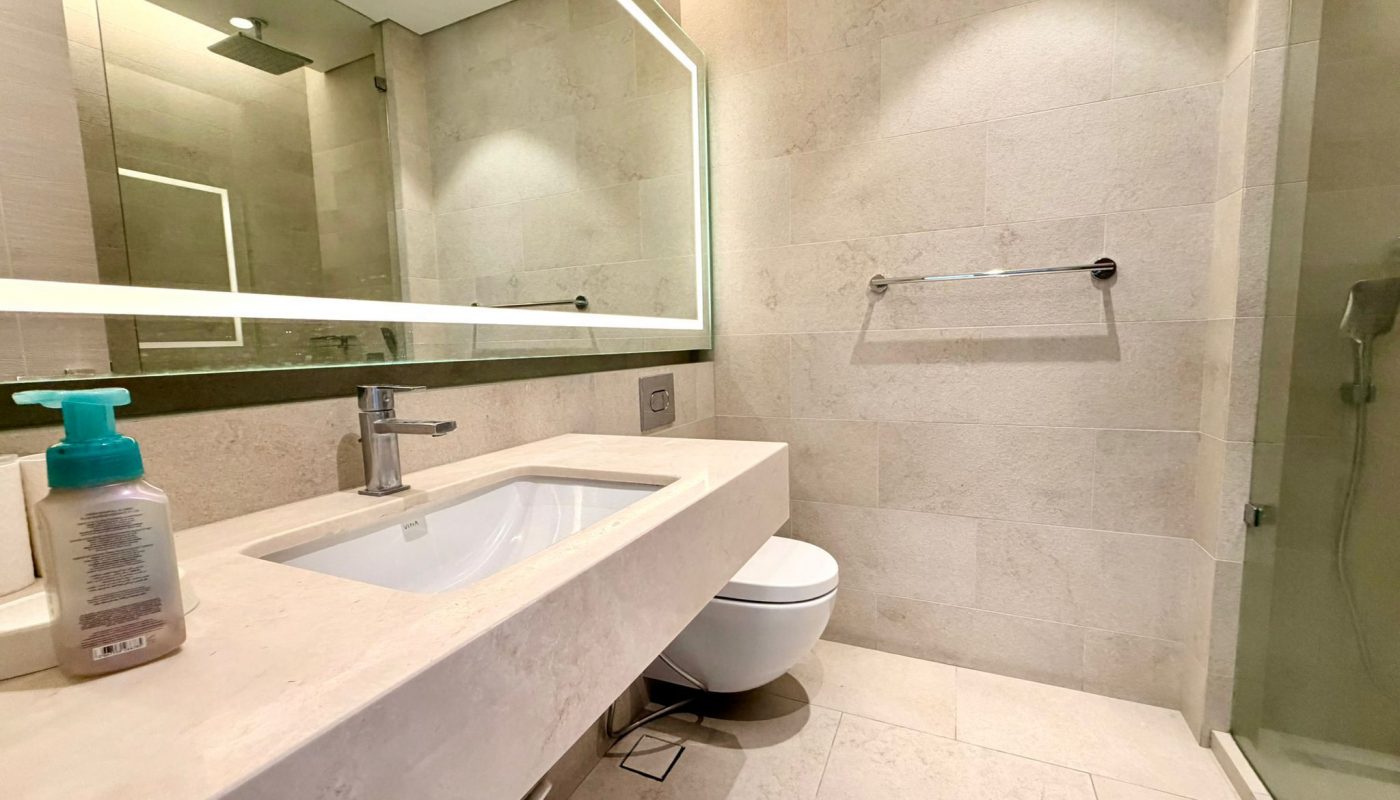 Modern bathroom with a large mirror, beige tiles, sink with soap dispenser, wall-mounted toilet, glass shower, and a towel rack.