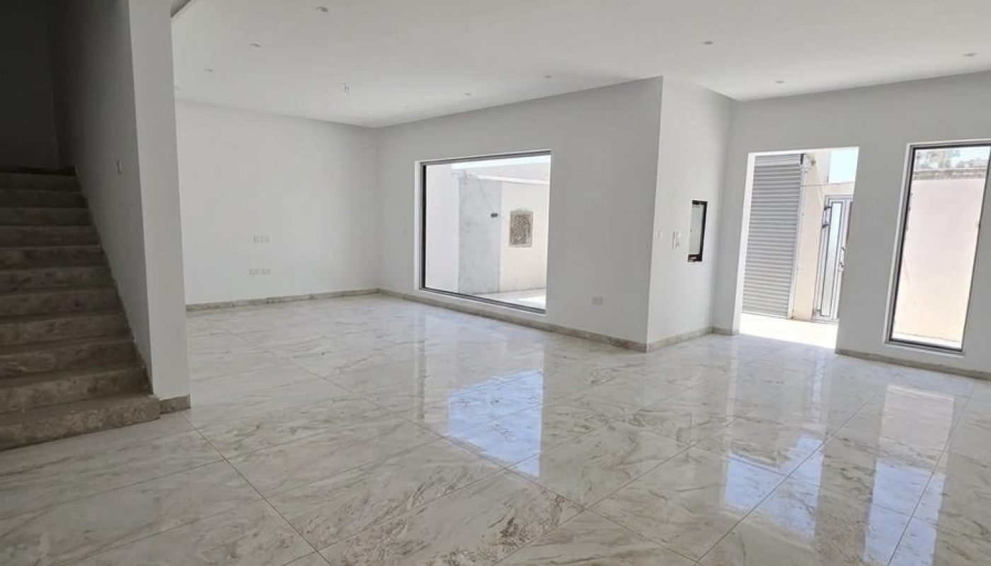 Empty, unfurnished room with marble tile floor, large windows, white walls, and a staircase leading to an upper floor. Natural light fills the space.