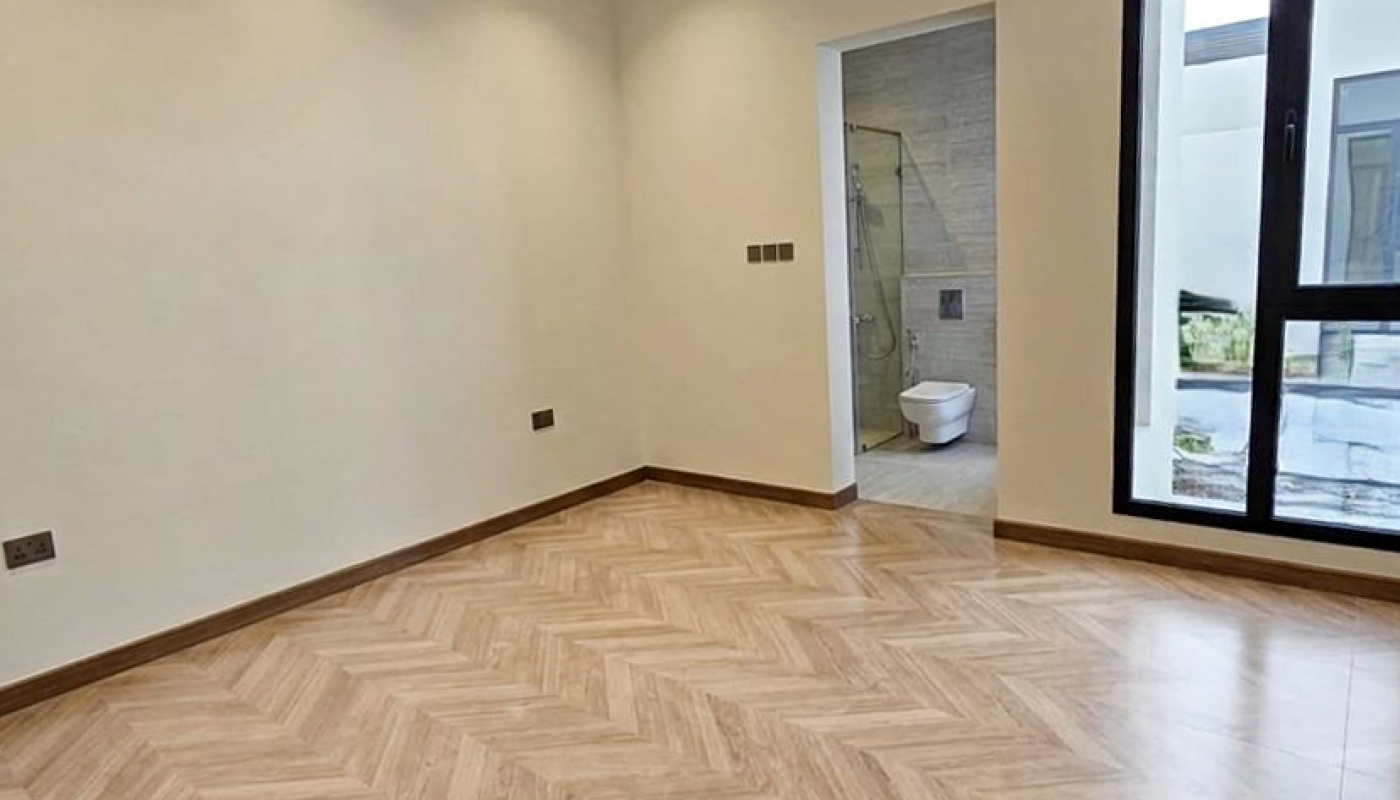 Empty room with light wood herringbone flooring, beige walls, recessed lighting, large window, and an open doorway leading to a bathroom with a visible toilet and shower.