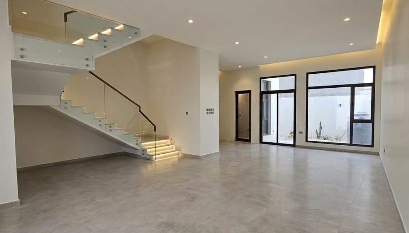Spacious modern living room with large windows, tiled floor, recessed lighting, and a floating glass staircase leading to an upper floor.