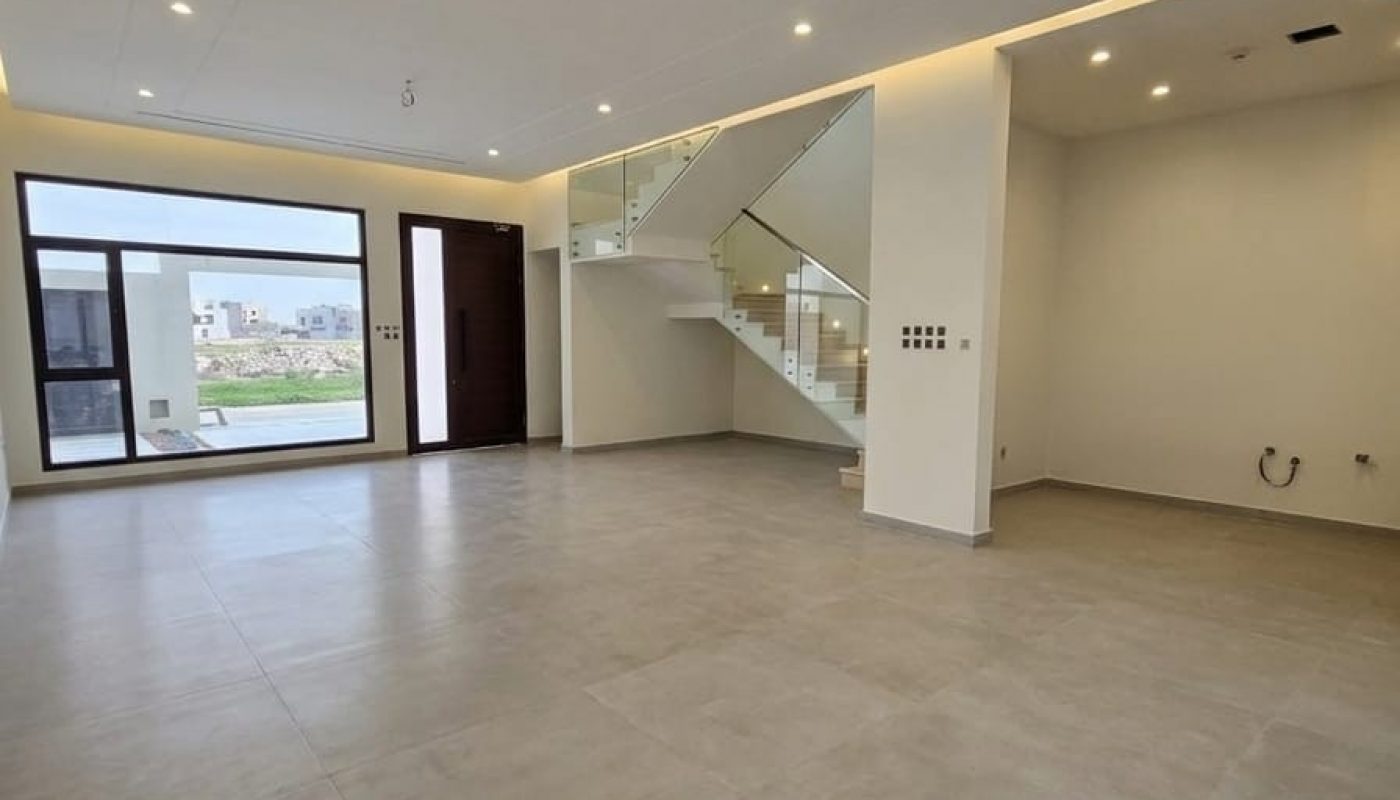 Spacious, modern empty room with large windows, tiled floors, recessed ceiling lights, and a glass railing staircase leading to an upper floor.