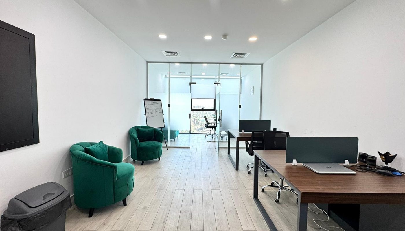 Modern office with two green chairs, desks with computers, a trash bin, a whiteboard, and a glass-walled meeting room at the back.