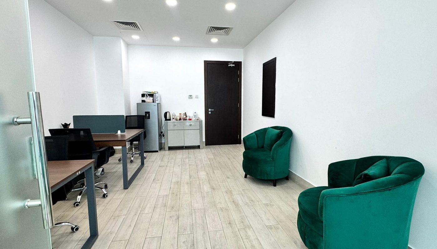 Modern office with two green armchairs, two work desks with chairs, a small cabinet, a coffee machine, and a mini refrigerator on a light wood floor.
