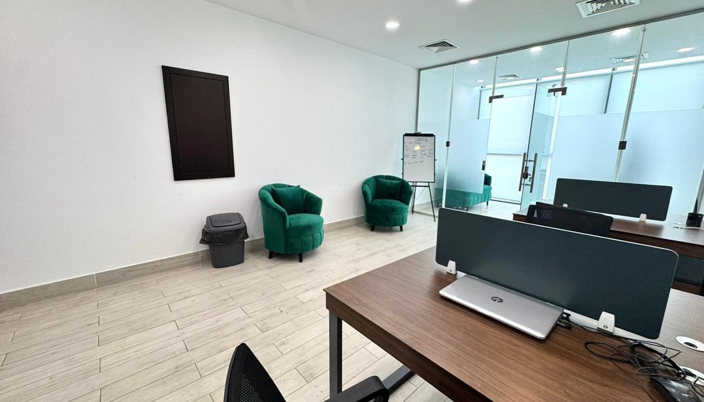 Modern office space with two green chairs, a trash bin, a flip chart, and desks with laptops near a glass partition.