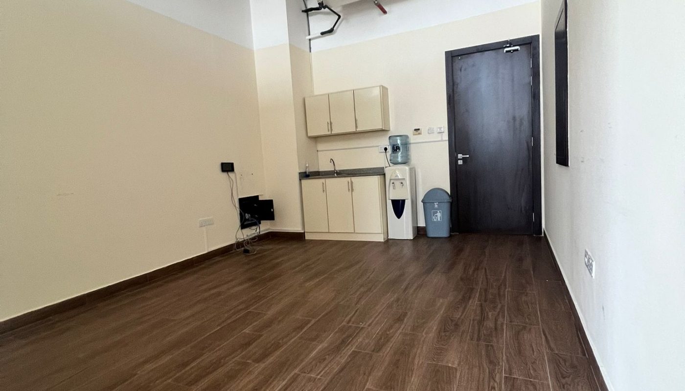 Empty room with wood-look flooring, beige walls, ceiling pipes, a kitchenette with cabinets, a water dispenser, a trash bin, and a black door at the far end.