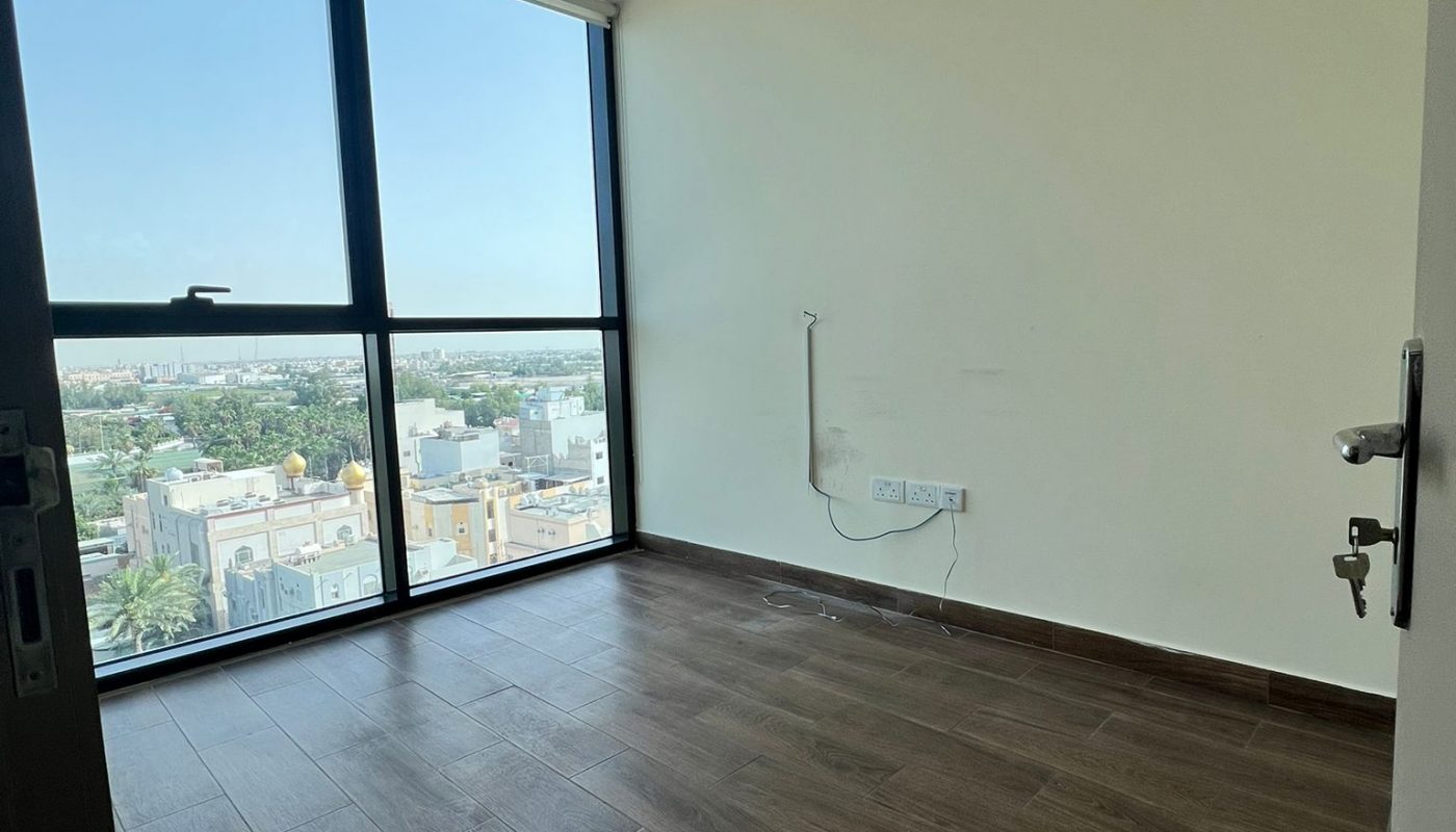 Empty room with large floor-to-ceiling window, brown wooden floor, cream walls, electrical outlets, and a city view outside.