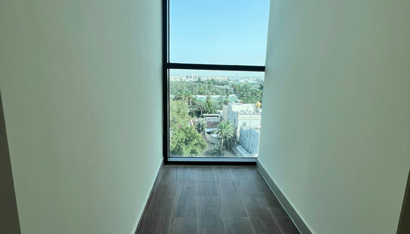 Narrow hallway with dark wood flooring leading to a large floor-to-ceiling window overlooking trees and buildings under a clear blue sky.