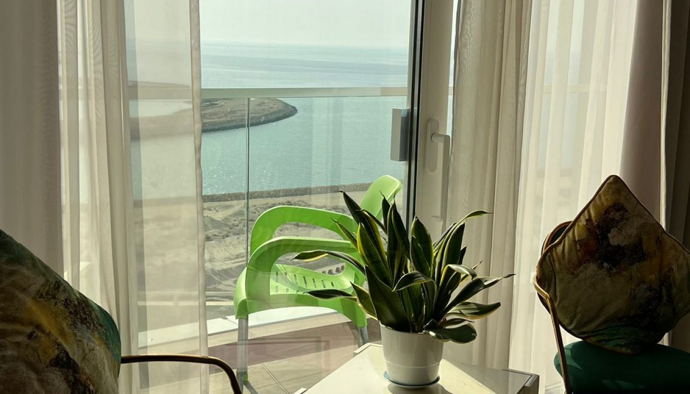 Two chairs and a small table with a potted plant are placed near a window with sheer curtains, overlooking a sunny beach and the sea.
