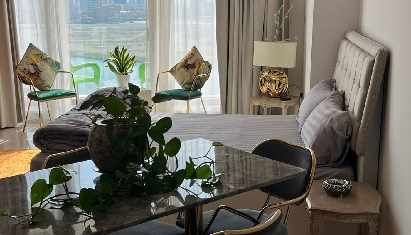 A modern living and dining area with a marble table, velvet chairs, orange rug, bed, decorative pillows, large windows, and a cityscape view in the background.