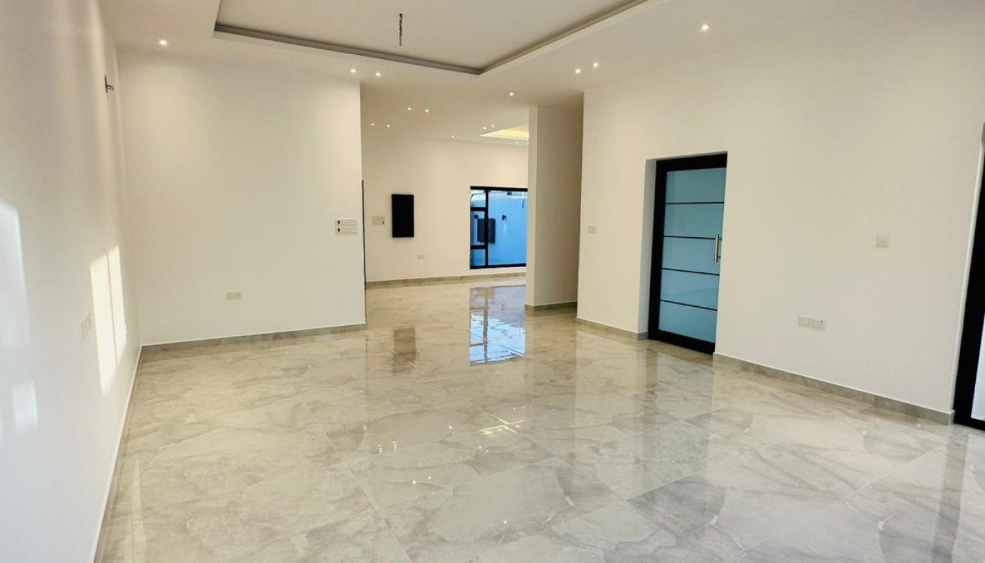 An empty, modern living room with white walls, a recessed ceiling with spotlights, and polished grey marble floors.