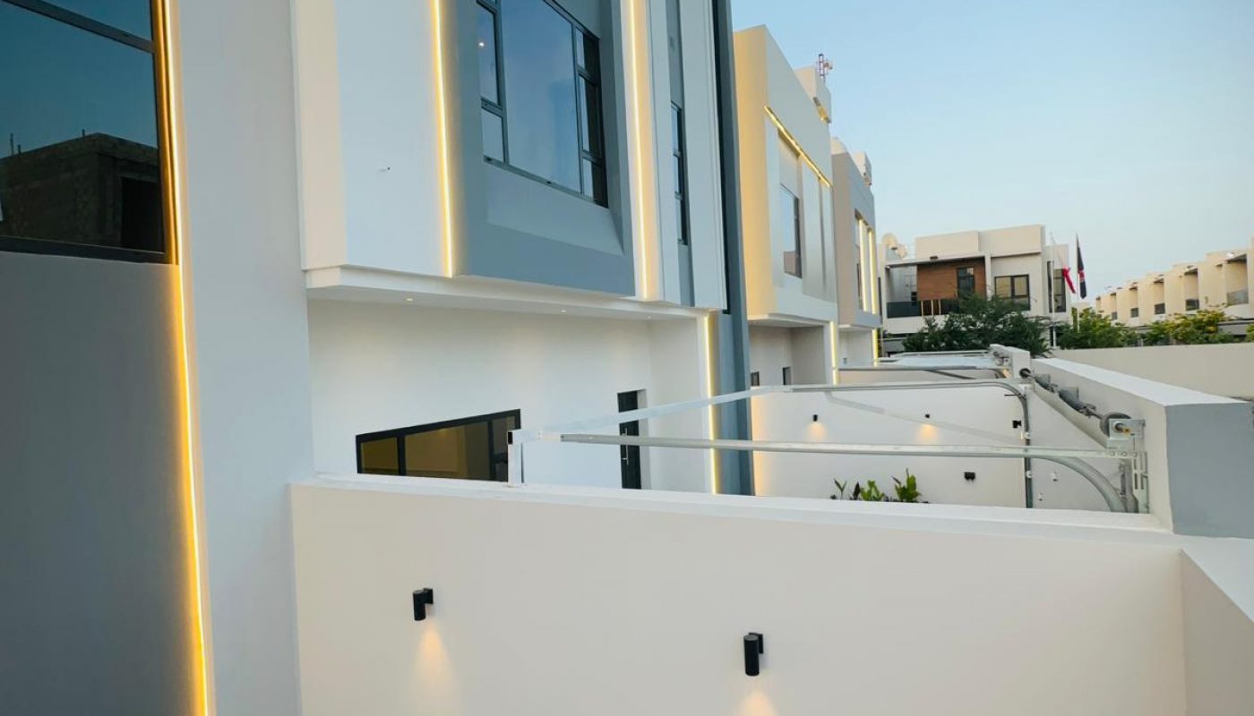 Modern townhouses with gray and white facades, vertical yellow lighting, and a small garden area with plants.