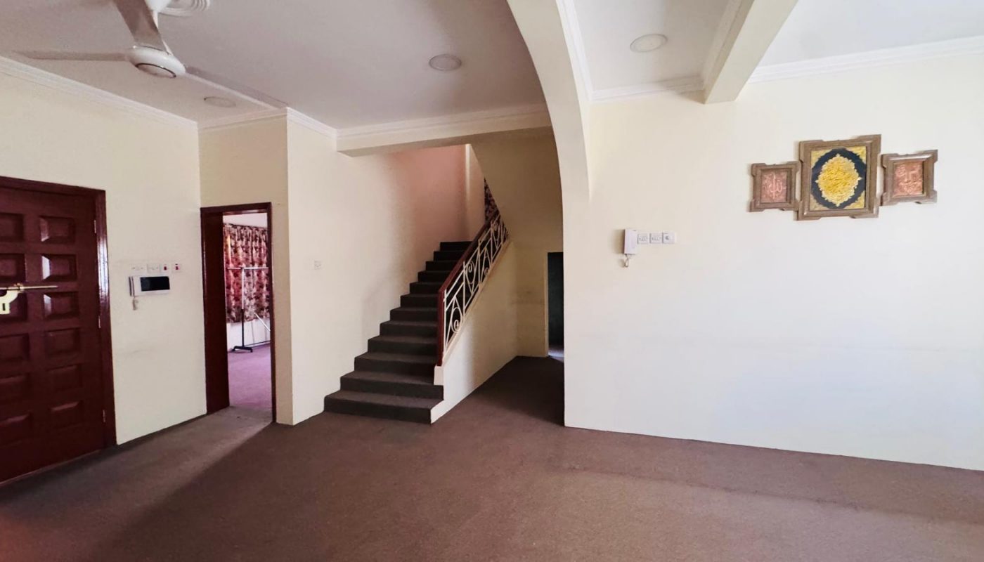 A room with carpeted floor, a staircase leading up, a ceiling fan, brown door on the left, and framed artwork on a cream wall.