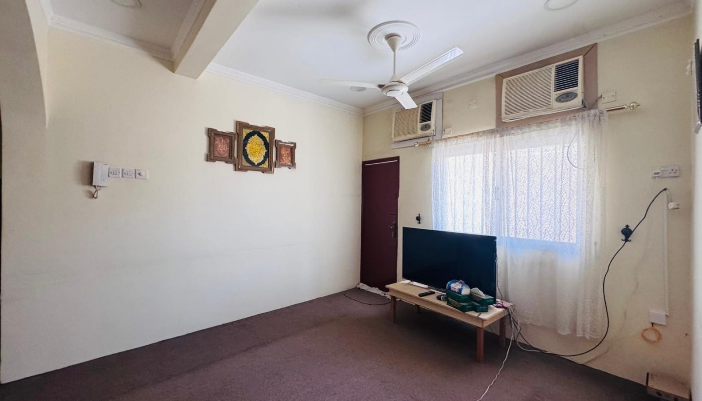 A living room with cream walls, a brown carpet, a ceiling fan, two wall-mounted air conditioners, a TV on a low table, and decorative items on the wall.
