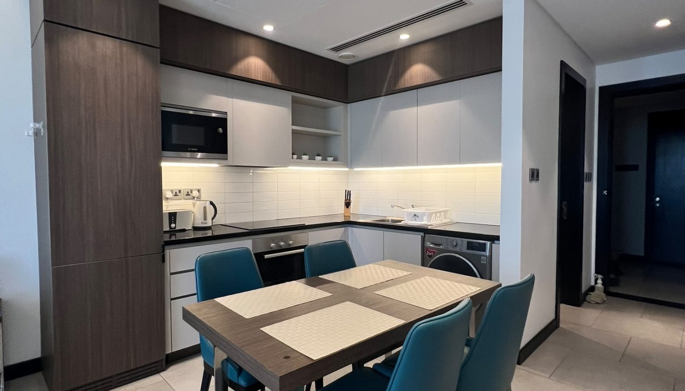 Modern kitchen with dark wood cabinets, white countertops, a dining table with four teal chairs, built-in appliances, and under-cabinet lighting.