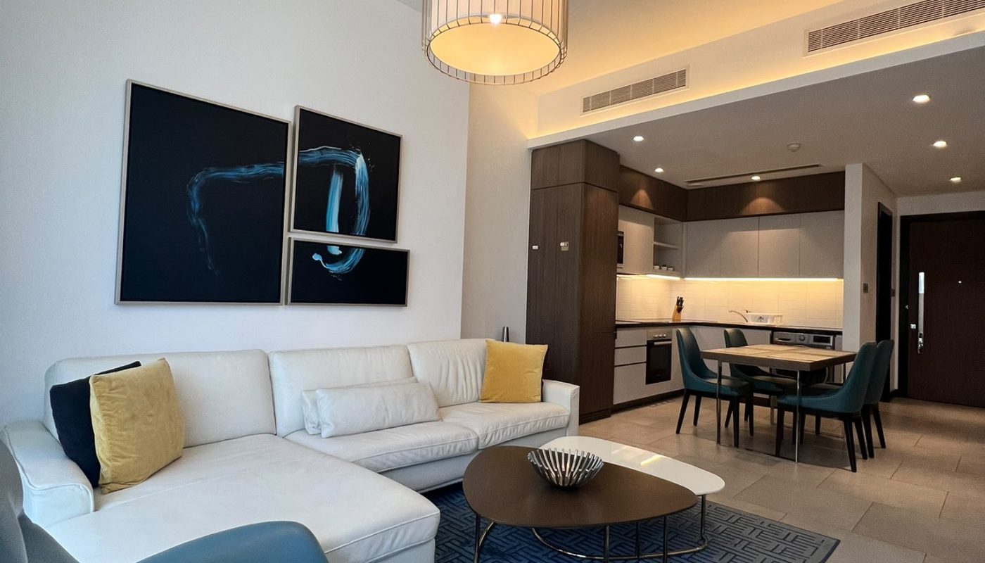 Modern living room with white sectional sofa, teal armchair, round coffee table, blue rug, wall art, and open kitchen in the background under two ceiling lights.