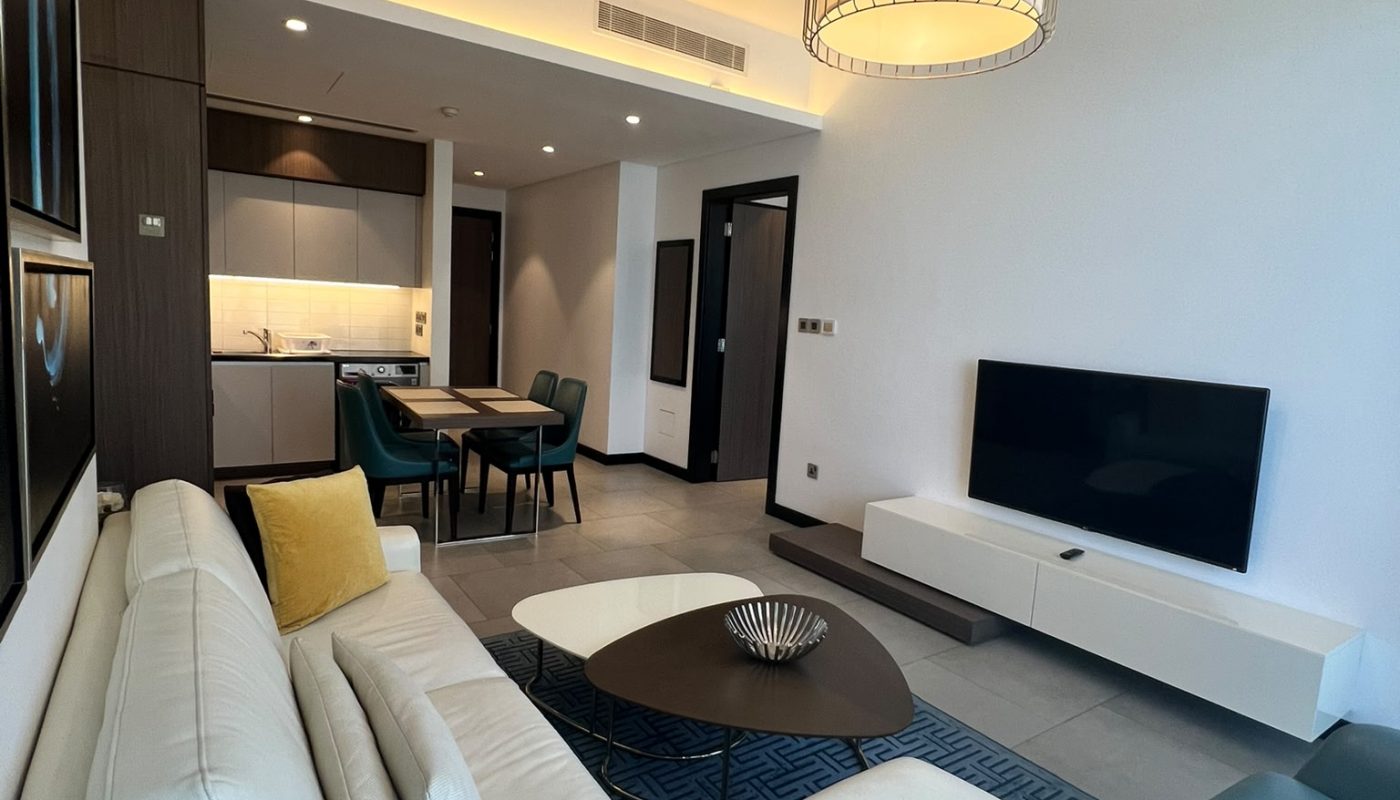 Modern living room with white sectional sofa, yellow cushion, round coffee table, wall-mounted TV, dining area, and open kitchen with sleek cabinets and minimalist decor.