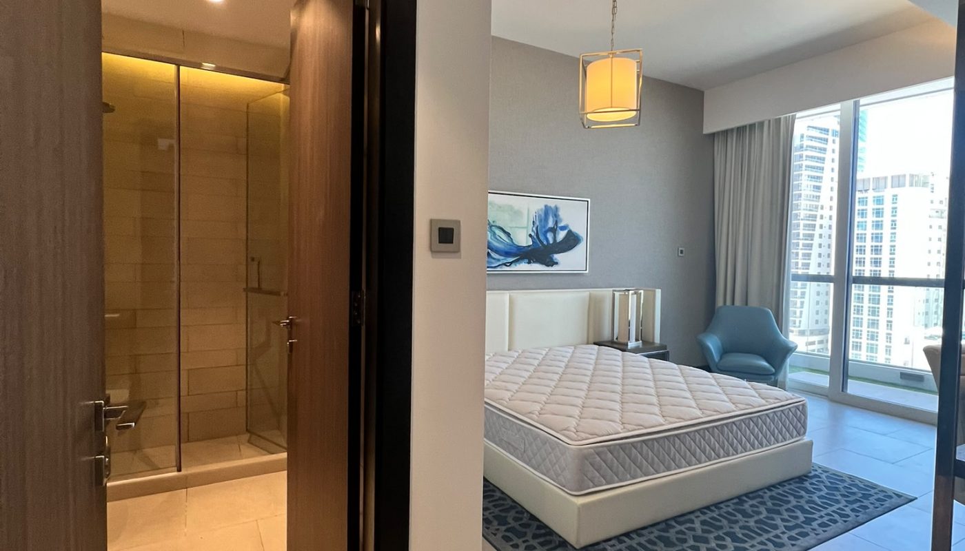 A modern bedroom with a large window, a double bed without bedding, a blue chair, wall art, and an open door leading to a bathroom with a glass shower.