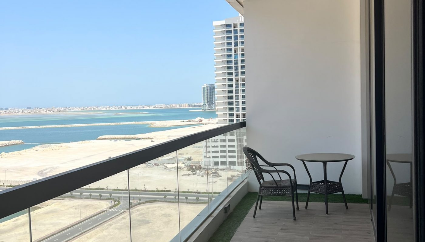 A modern balcony with glass railing, two chairs, and a table overlooks a sandy area, water, and buildings in the distance under a clear blue sky.