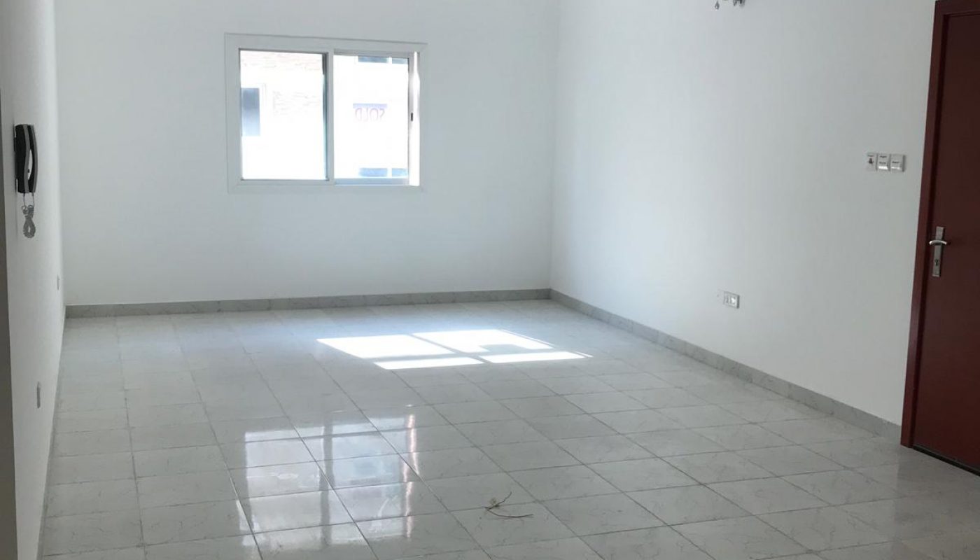 Empty, unfurnished room with white walls, tiled floor, a window, a red door, ceiling lights, and wall fixtures. Sunlight shines through the window onto the floor.