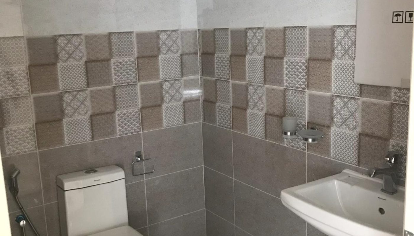 A bathroom with a toilet, bidet sprayer, pedestal sink, soap dispenser, and tiled walls in a geometric pattern.