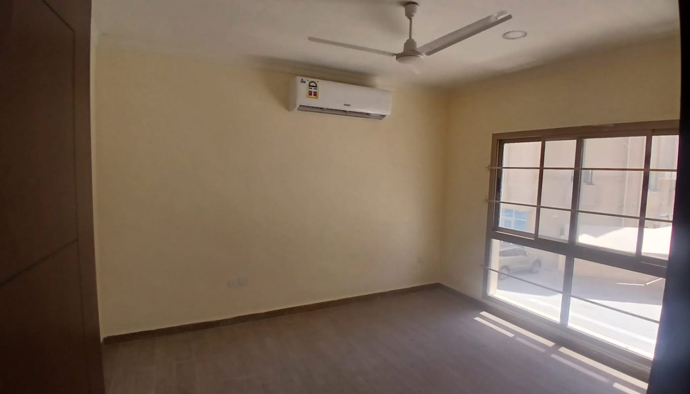 Empty room with beige walls, tiled flooring, ceiling fan, split air conditioning unit, and a large window letting in natural light.