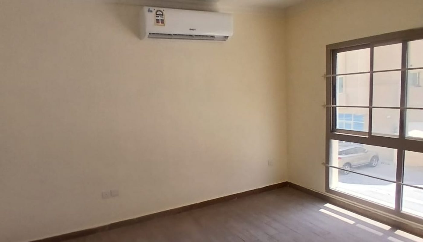 Empty room with beige walls, wooden floor, a ceiling fan, air conditioner, and a large window letting in sunlight.