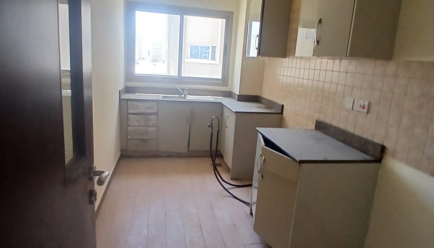 A small, unfurnished kitchen with light wood flooring, tiled backsplash, grey cabinets, and a window above the sink letting in natural light.