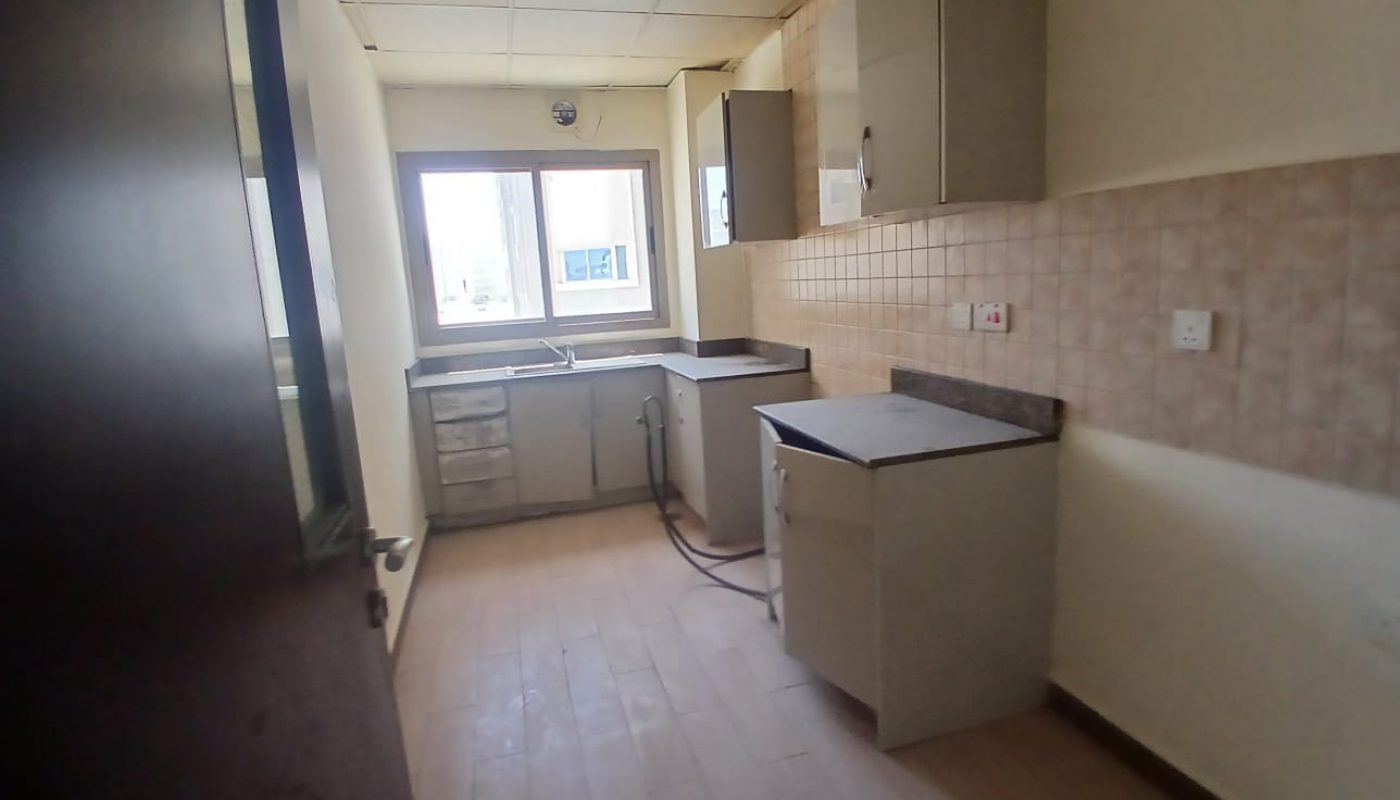 Narrow kitchen with light-colored cabinets, a black countertop, tiled backsplash, and a window above the sink; ceiling tiles and light wooden flooring are visible.