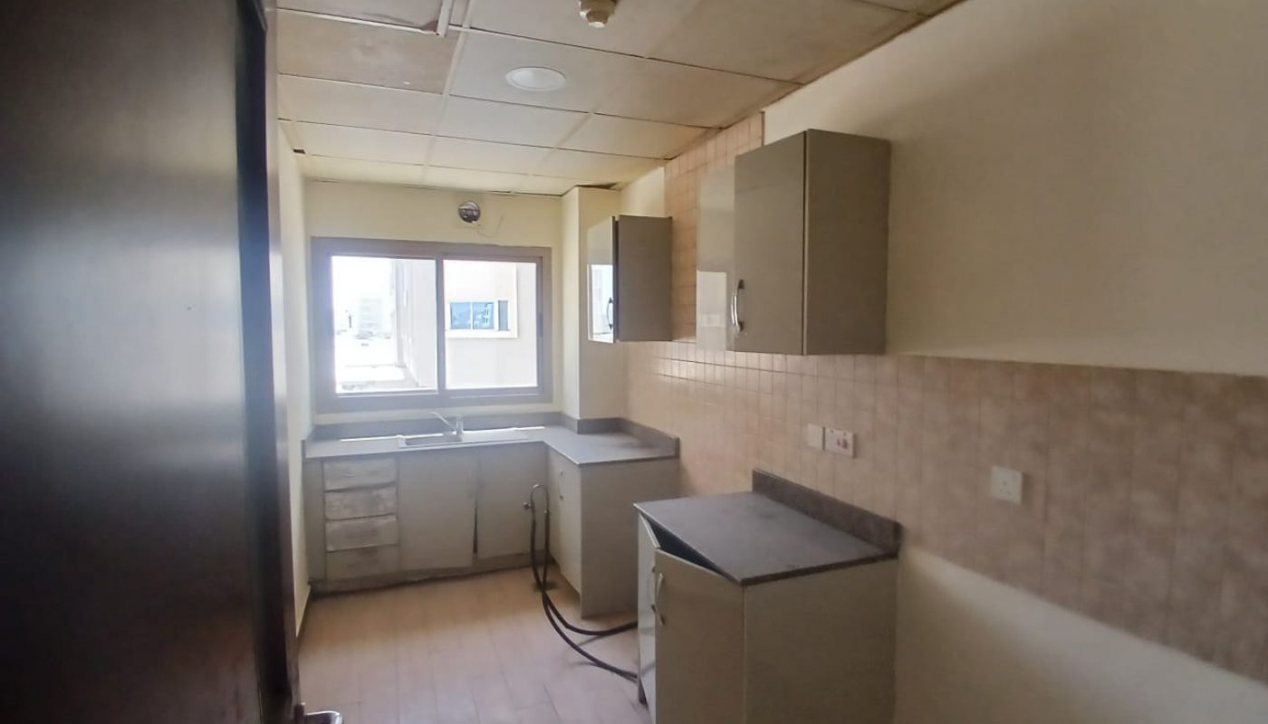 A small, empty kitchen with tiled walls, overhead cabinets, a window above the sink, and basic countertops and storage units.