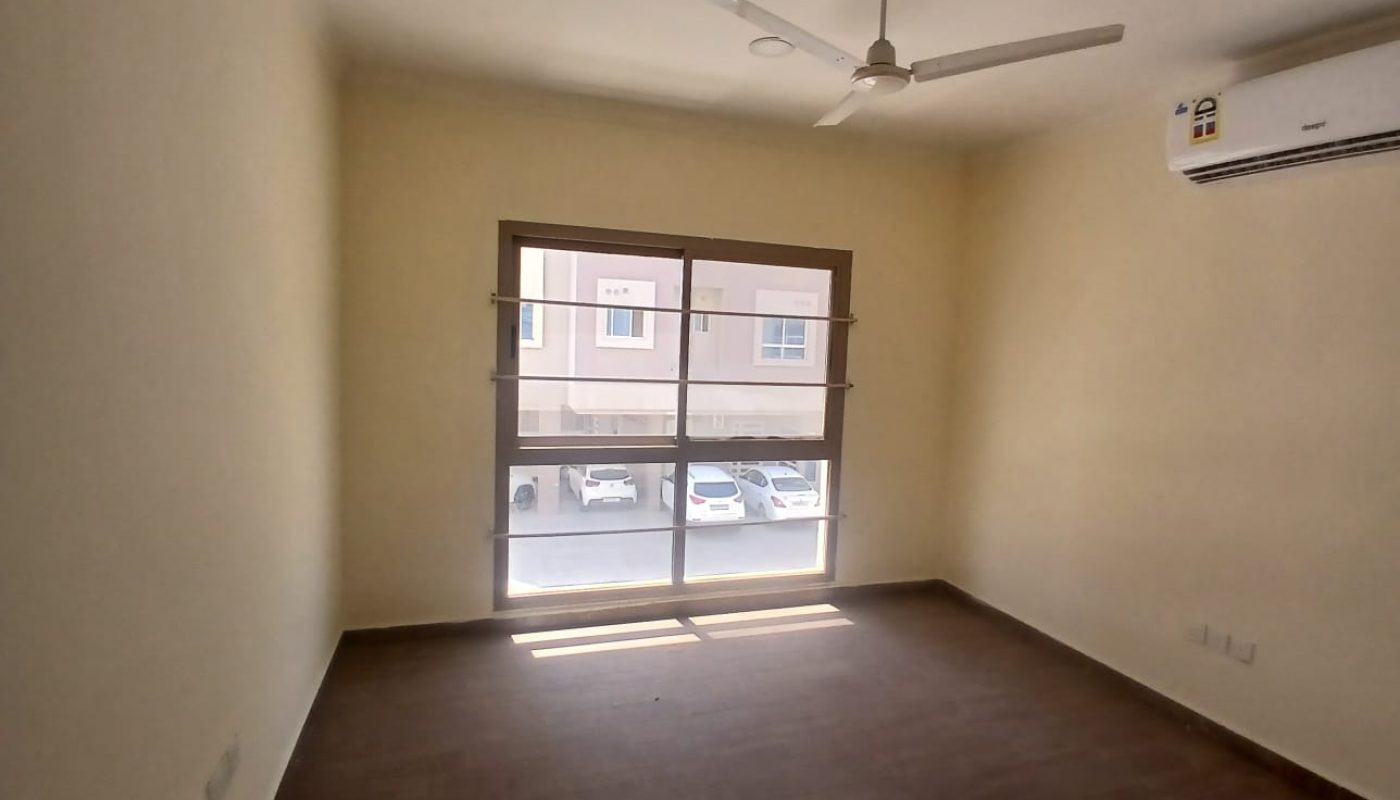 Empty room with brown flooring, beige walls, a large window, ceiling fan, and wall-mounted air conditioner. Several parked cars are visible outside the window.