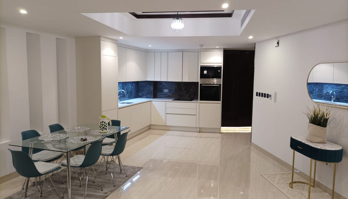 Modern open-plan kitchen and dining area with white cabinets, blue chairs around a glass table, marble flooring, and a small side table with a plant.