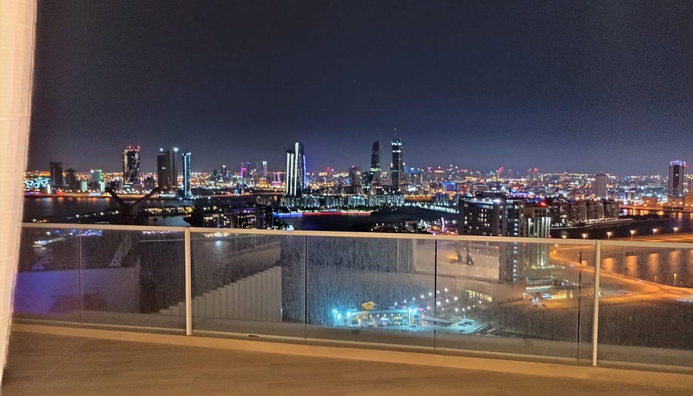 Night view of a city skyline with illuminated buildings and a river, seen from a balcony with glass railing and wooden floor.