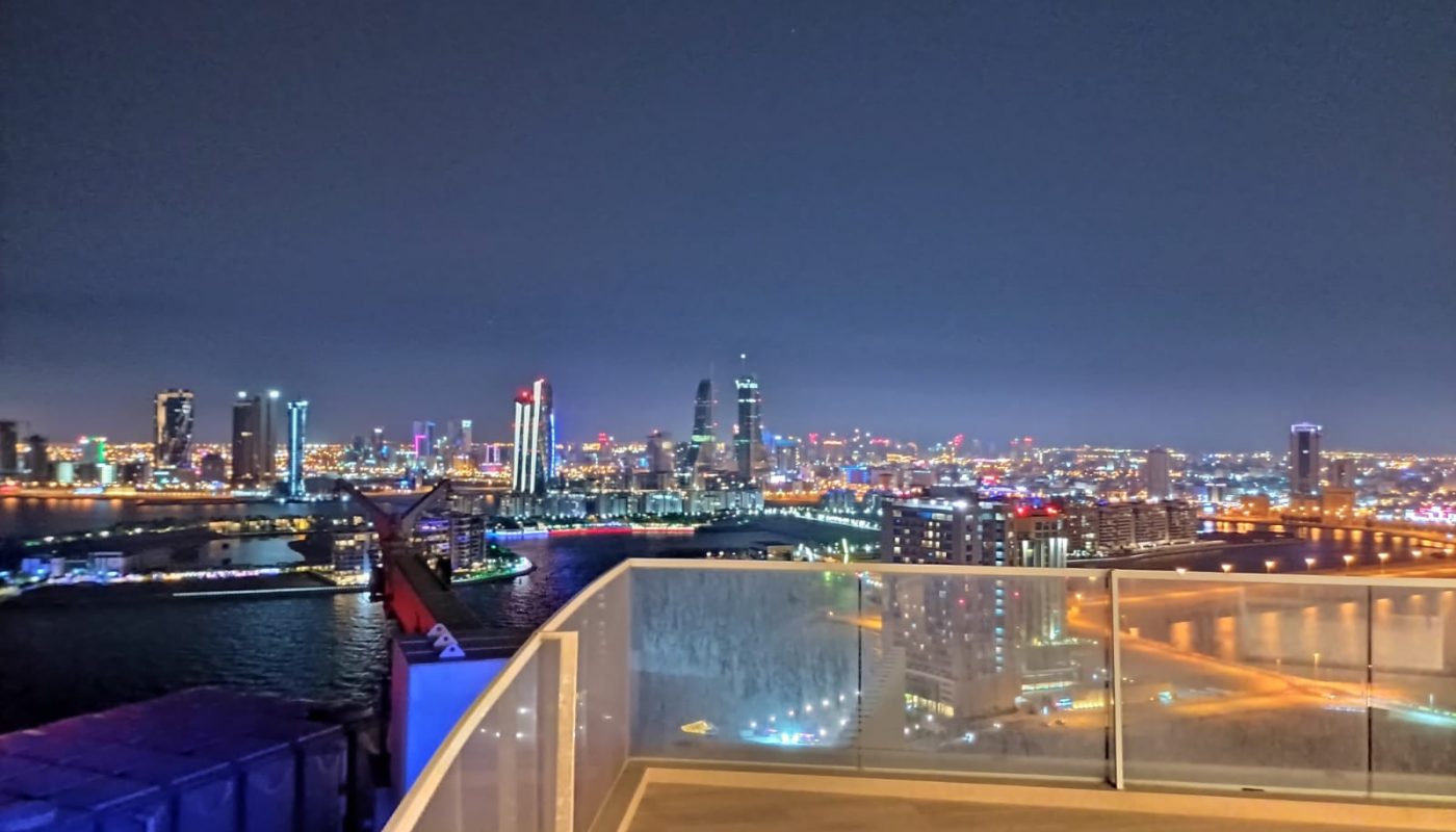Nighttime cityscape view from a rooftop with glass railings, overlooking a river, illuminated buildings, and a skyline with colorful lights.