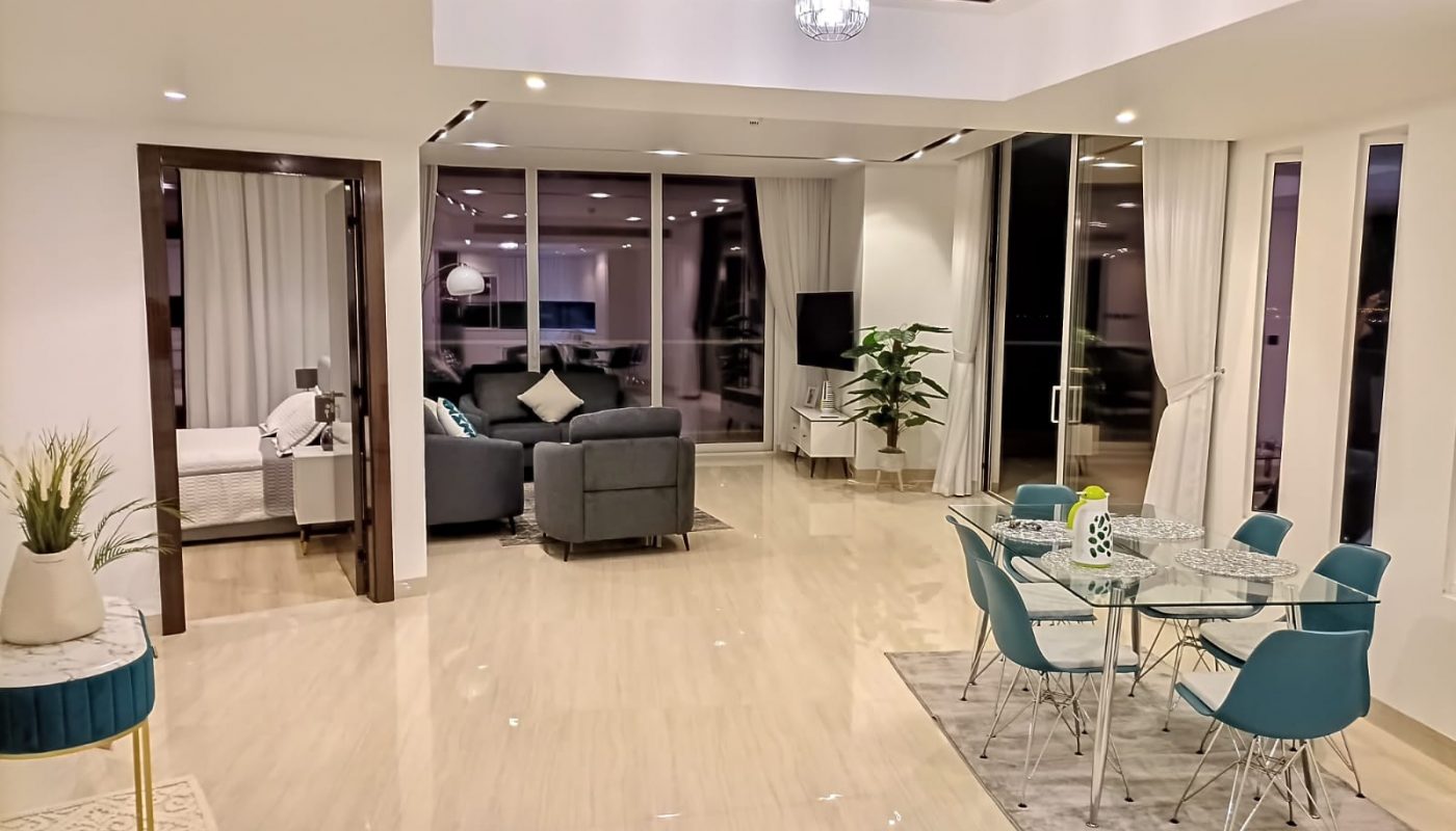 Modern open-plan living space with dining area, glass table and teal chairs, adjacent to a seating area and a bedroom, featuring large windows and neutral decor.