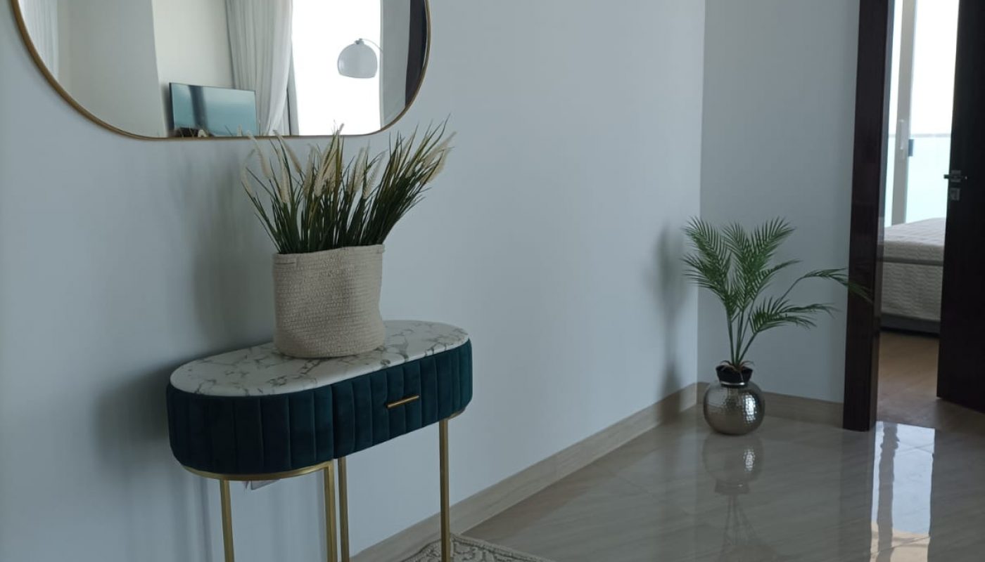 A modern hallway with a blue console table, a potted plant, a decorative rug, an oval mirror on the wall, and another potted plant near an open doorway leading to another room.
