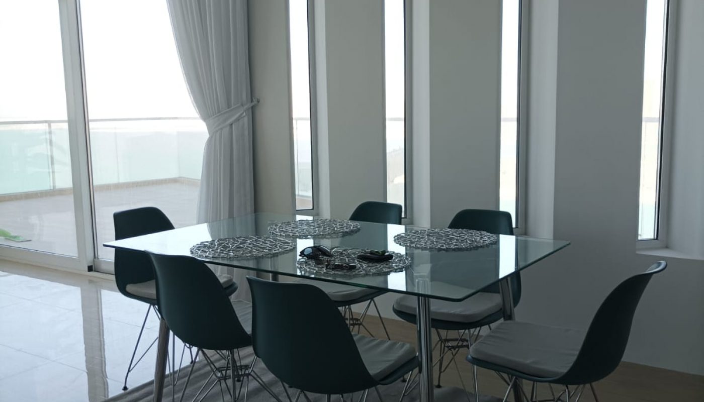 A modern dining area with a glass table, six black chairs, a grey rug, and tall windows letting in natural light.