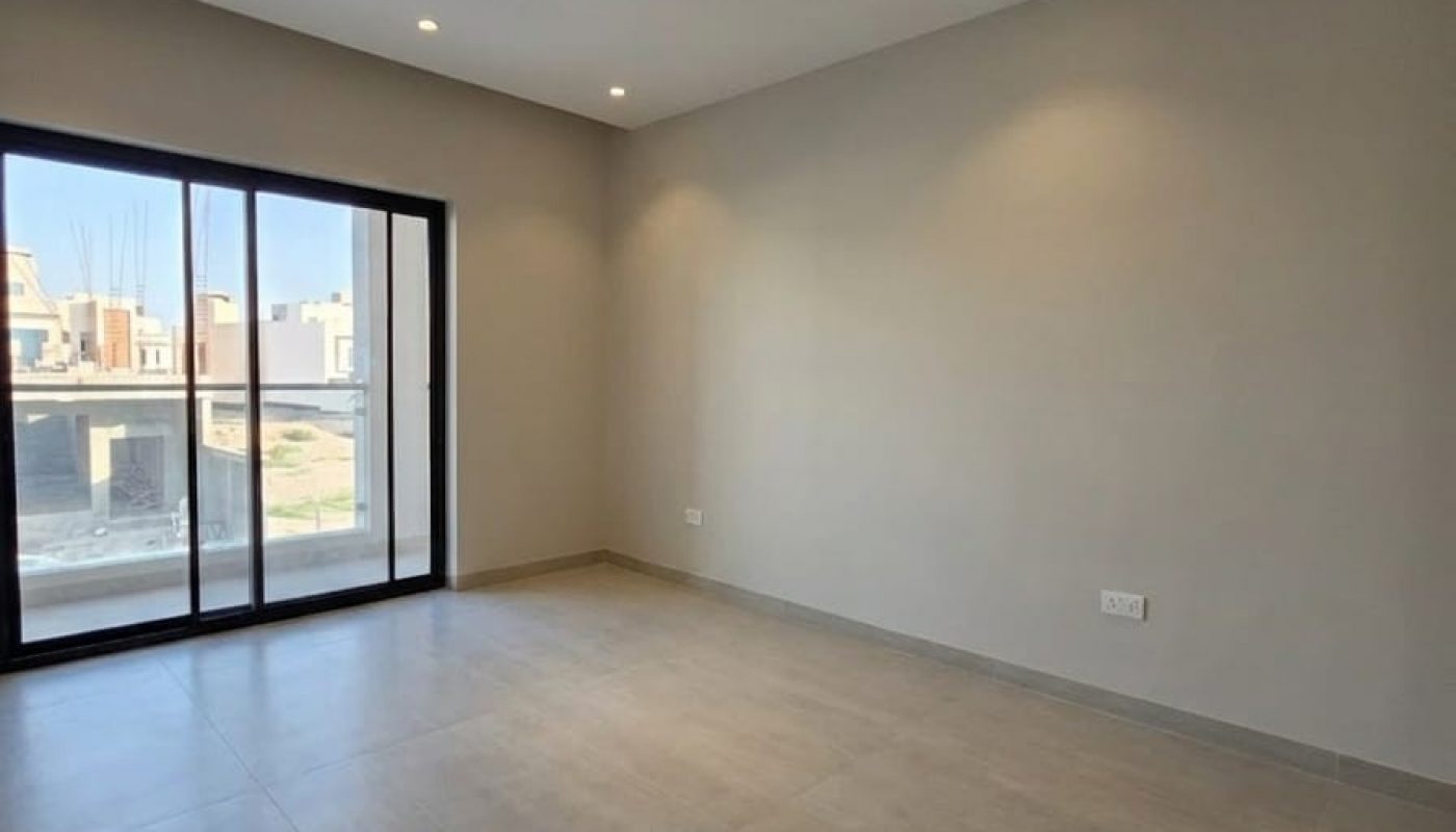 Empty room with beige tiled floor, plain light-colored walls, recessed ceiling lights, and large sliding glass doors leading to a balcony with an outdoor view.