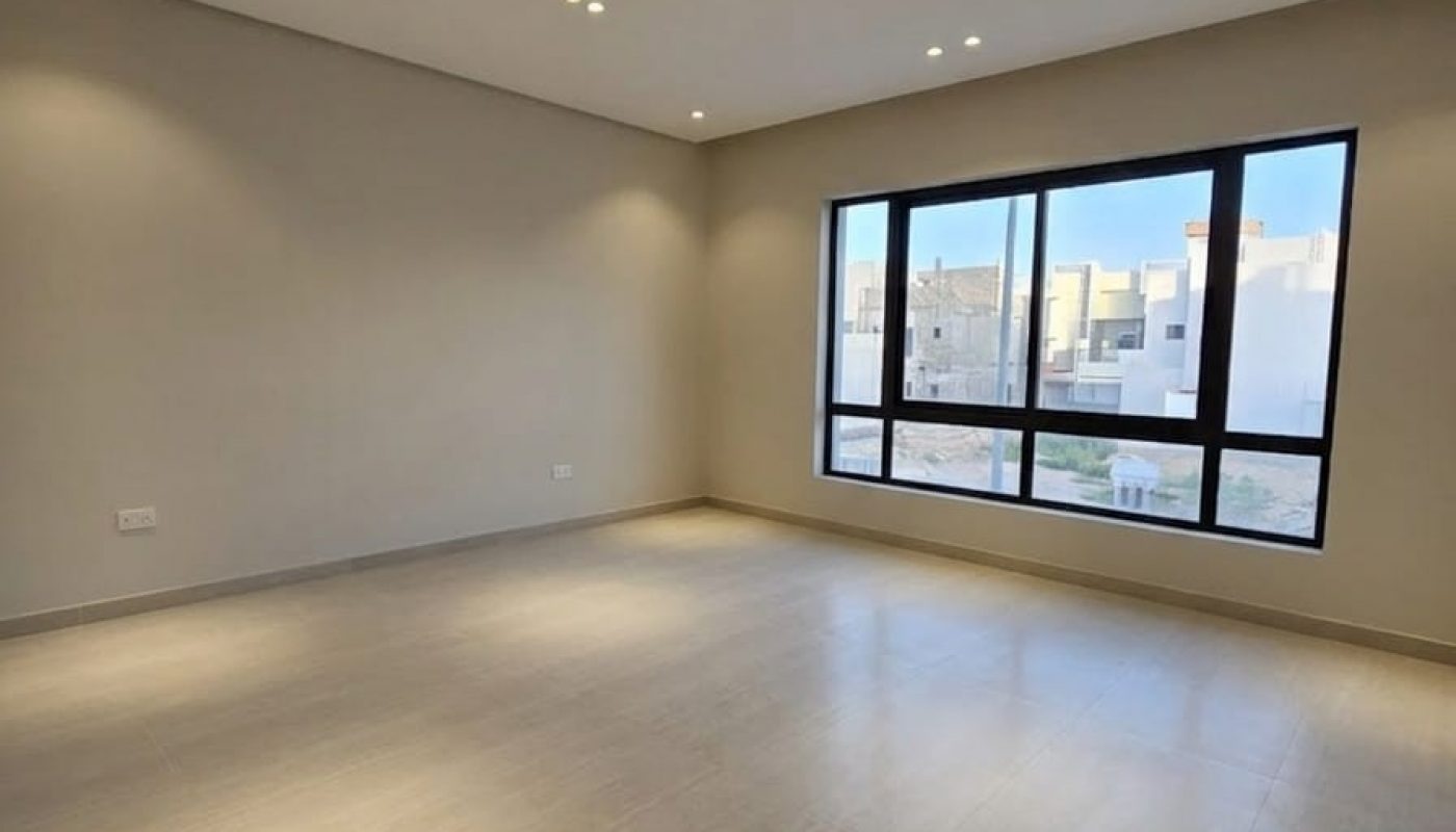An empty room with light-colored tiled flooring, beige walls, recessed ceiling lights, and a large window showing buildings outside.