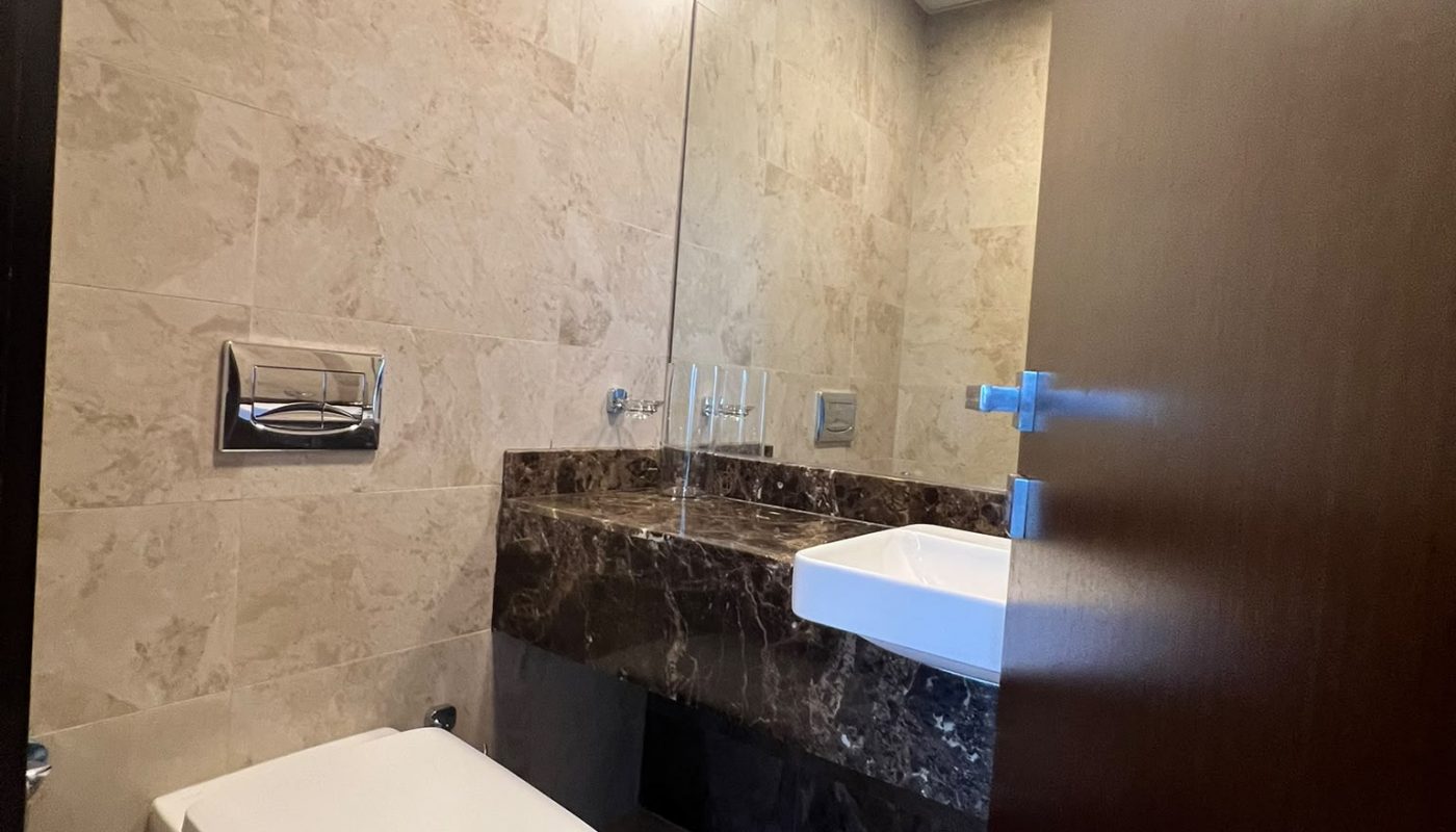 Modern bathroom with beige marble walls and floor, wall-mounted toilet, large mirror, dark marble countertop, and white sink. Door is partially open.