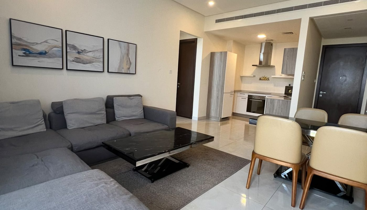 Modern living room and kitchen with gray sectional sofa, glass coffee table, beige dining chairs, wall art, and tile flooring.