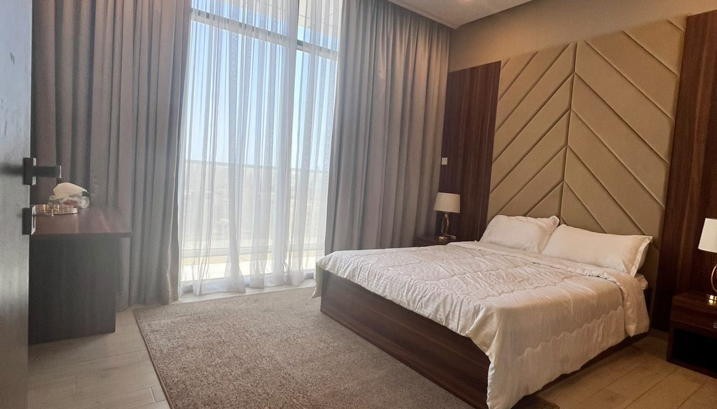 Modern bedroom with a large bed, wooden floor, area rug, two nightstands with lamps, and tall windows covered by sheer and blackout curtains.