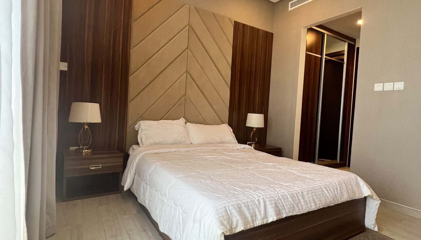 A modern bedroom with a double bed, white bedding, wooden headboard, two bedside tables with lamps, a large rug, and mirrored closet doors.