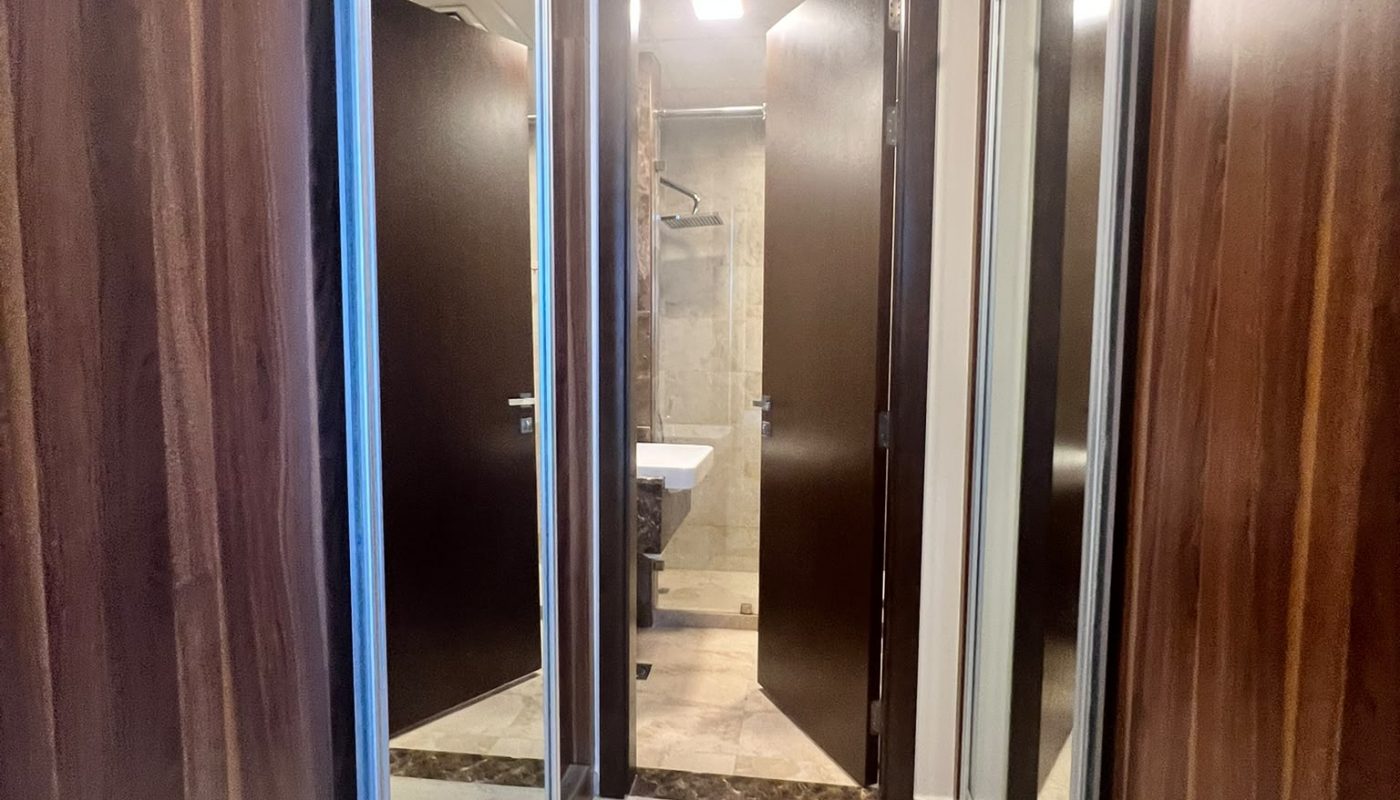 A narrow hallway with wooden closet doors on both sides leads to an open door revealing a bathroom with beige tiles and a sink.