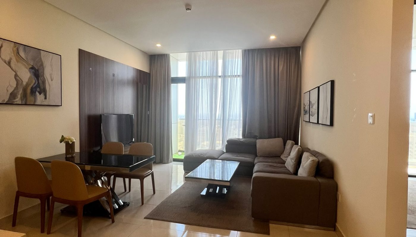 A modern living room with a large sectional sofa, dining table with chairs, TV, wall art, tiled floor, and floor-to-ceiling windows with sheer curtains.