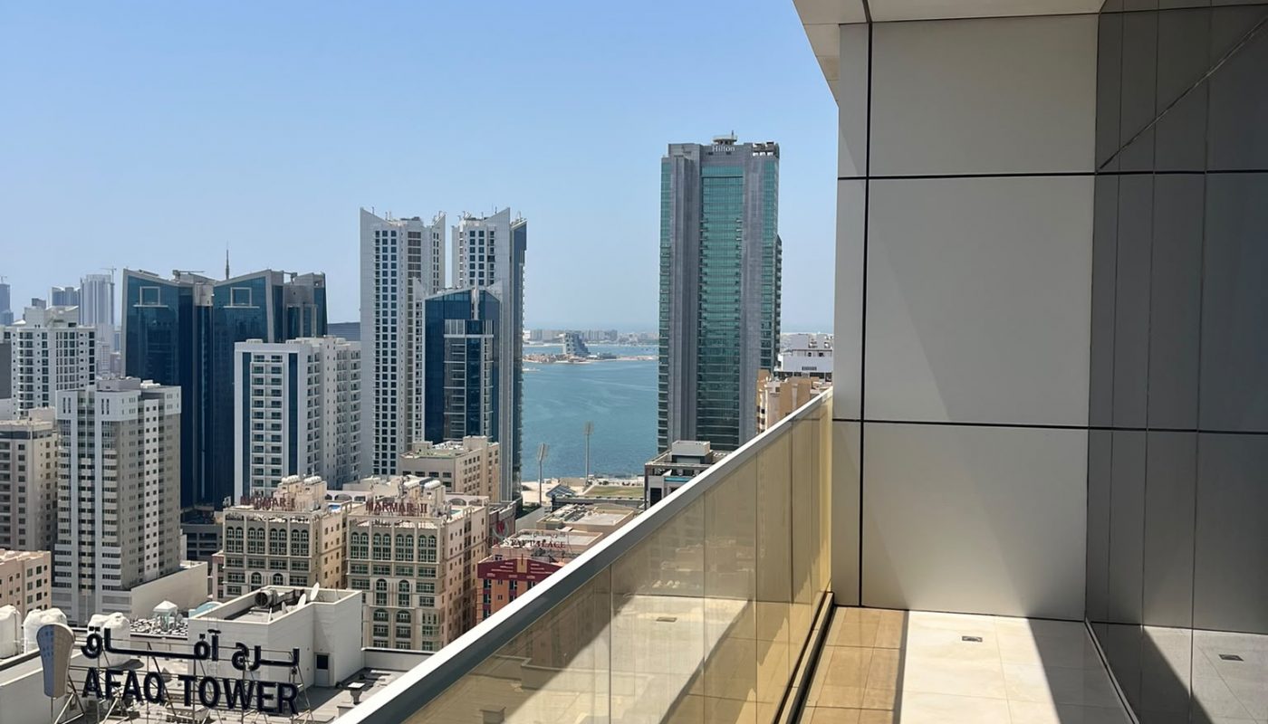 High-rise balcony with glass railing overlooking city buildings and water under a clear blue sky. Shadows fall across the tiled floor.
