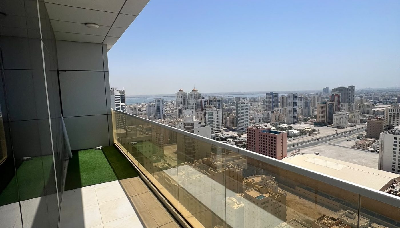High-rise balcony with glass railing overlooks a cityscape with tall buildings under a clear blue sky on a sunny day.