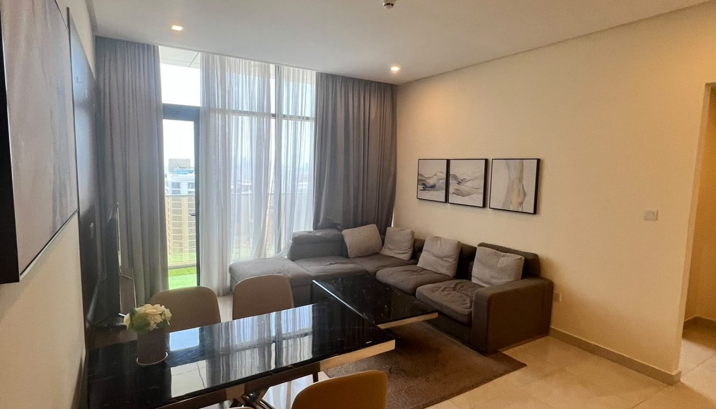 A modern living and dining area with a glass-top table, four chairs, an L-shaped sofa, wall art, large windows with sheer curtains, and tiled floors.