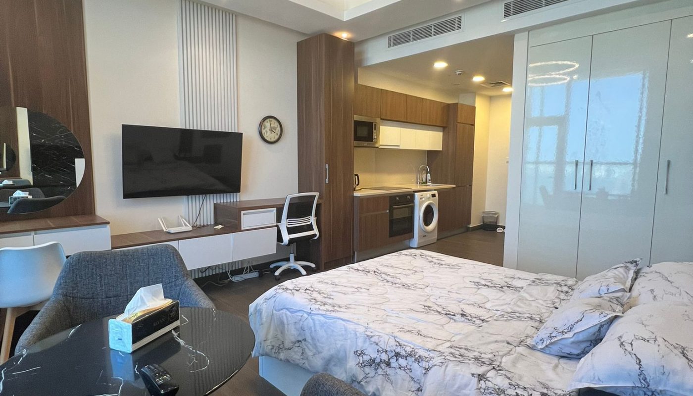 Modern studio apartment with a bed, small dining table, TV, desk, and kitchenette; neutral tones and contemporary lighting.