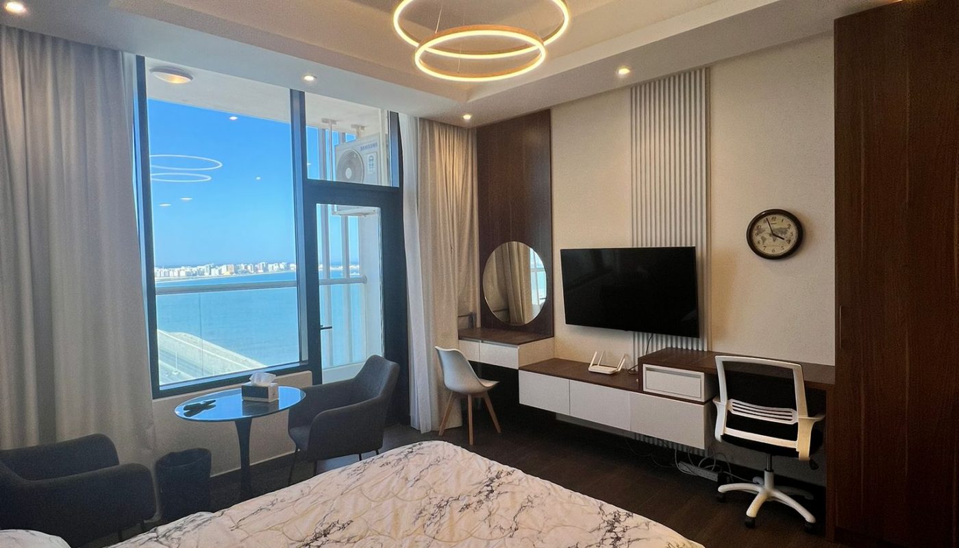 Modern bedroom with marble-pattern bedding, desk, wall-mounted TV, two chairs by a window with city view, and circular ceiling lights.